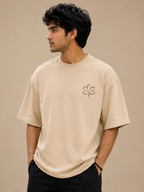 A man wearing a beige crewneck t-shirt with a small black line-art lotus flower graphic on the left chest, paired with black pants, set against a solid beige background.