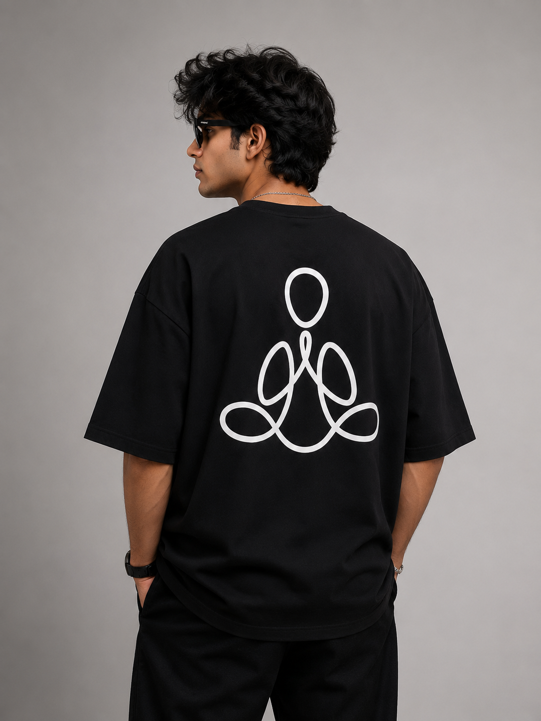 A man wearing a black oversized t-shirt with a white continuous line graphic on the back, shown from behind against a neutral gray studio background.