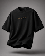 A black oversized short-sleeve t-shirt with the word 'PEACE' printed in gold across the chest, shown floating against a neutral gray background.