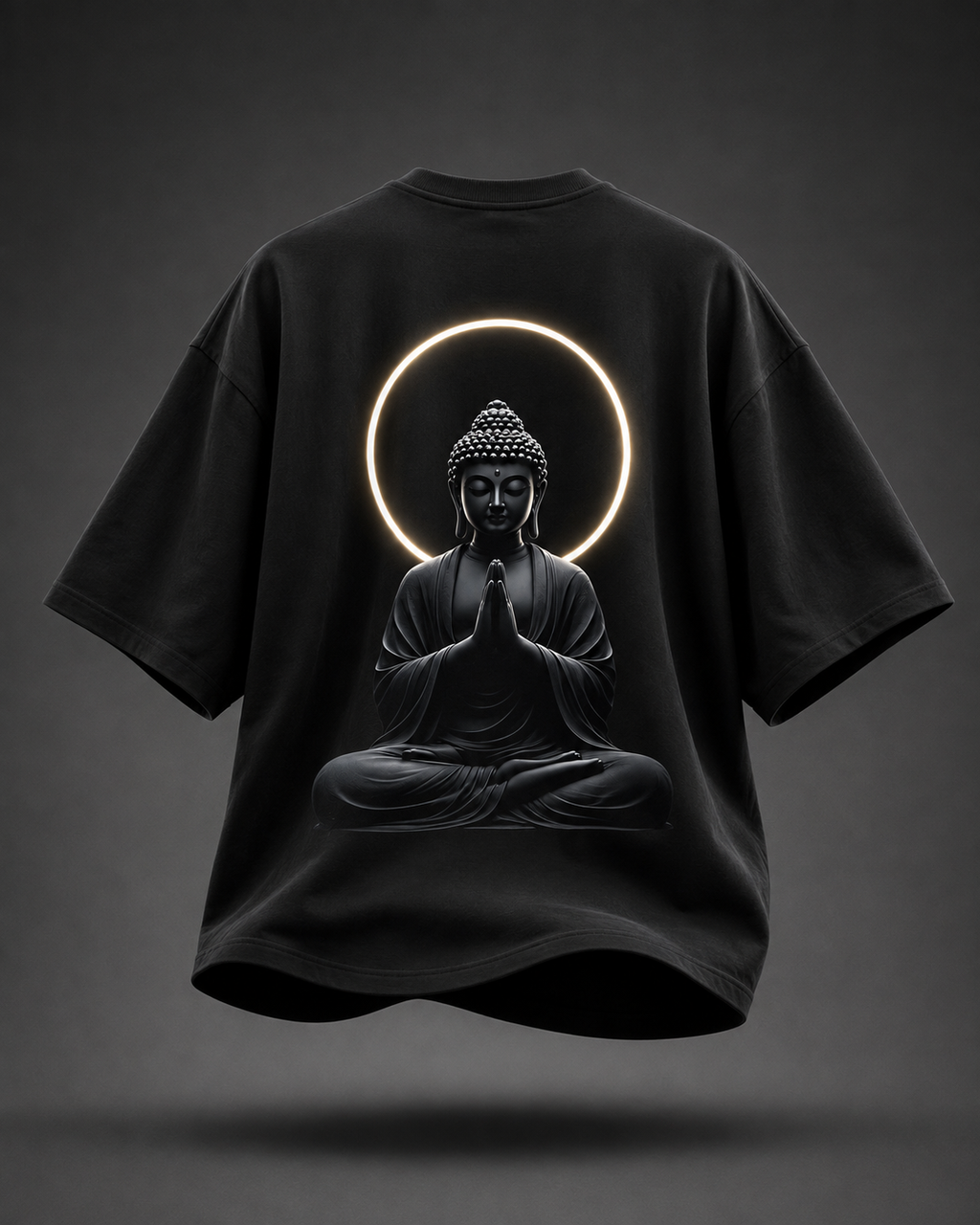 A black oversized short-sleeve T-shirt shown from the back, featuring a large graphic of a seated Buddha figure with hands in prayer, framed by a glowing circular halo. The shirt is centered on a dark gray gradient background with a soft shadow beneath it.