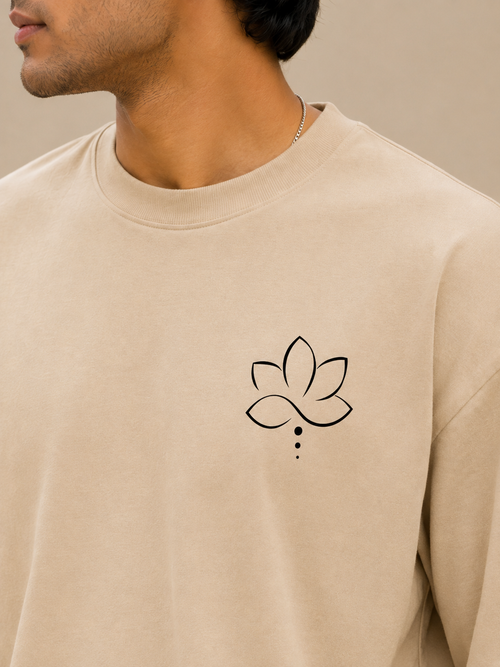 A close-up of a man wearing a beige crewneck sweatshirt with a black line-art lotus flower graphic on the left chest.
