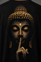 A close-up of a black t-shirt featuring a detailed graphic print of a Buddha face in gold and bronze tones, with the figure's finger resting on its lips in a shushing gesture.