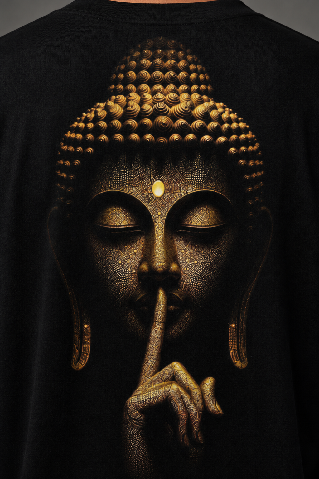 A close-up of a black t-shirt featuring a detailed graphic print of a Buddha face in gold and bronze tones, with the figure's finger resting on its lips in a shushing gesture.