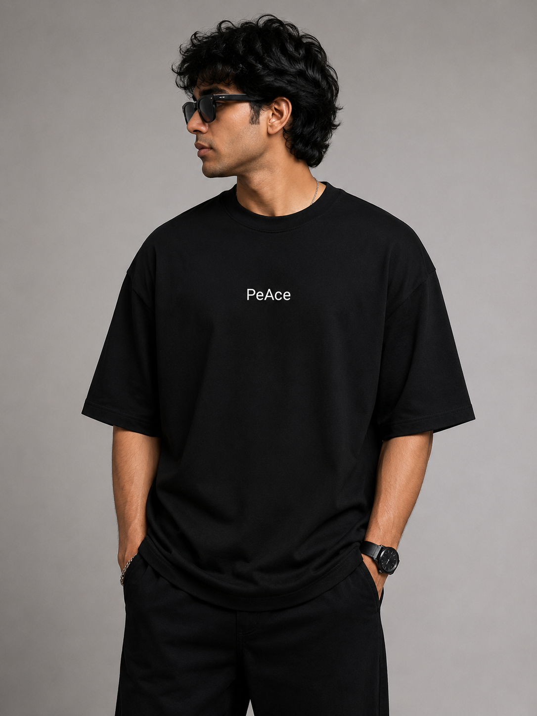 A man wearing a black oversized t-shirt with the word 'PeAce' printed in white on the chest, paired with black pants, set against a solid gray studio background.