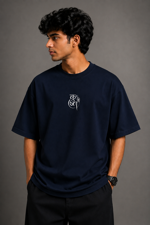 A man wearing a navy blue crewneck t-shirt with a white stylized graphic on the left chest, paired with black pants, standing against a dark gray studio background.